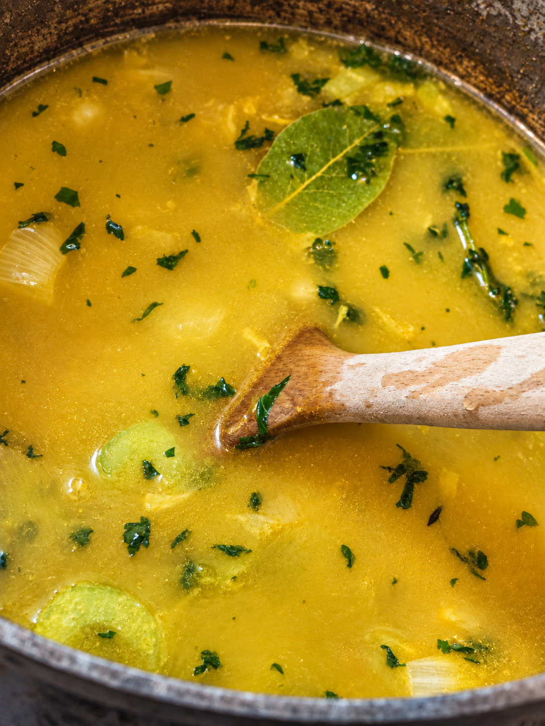 Golden chickpea noodle soup broth with vegetables, herbs, and a wooden spoon