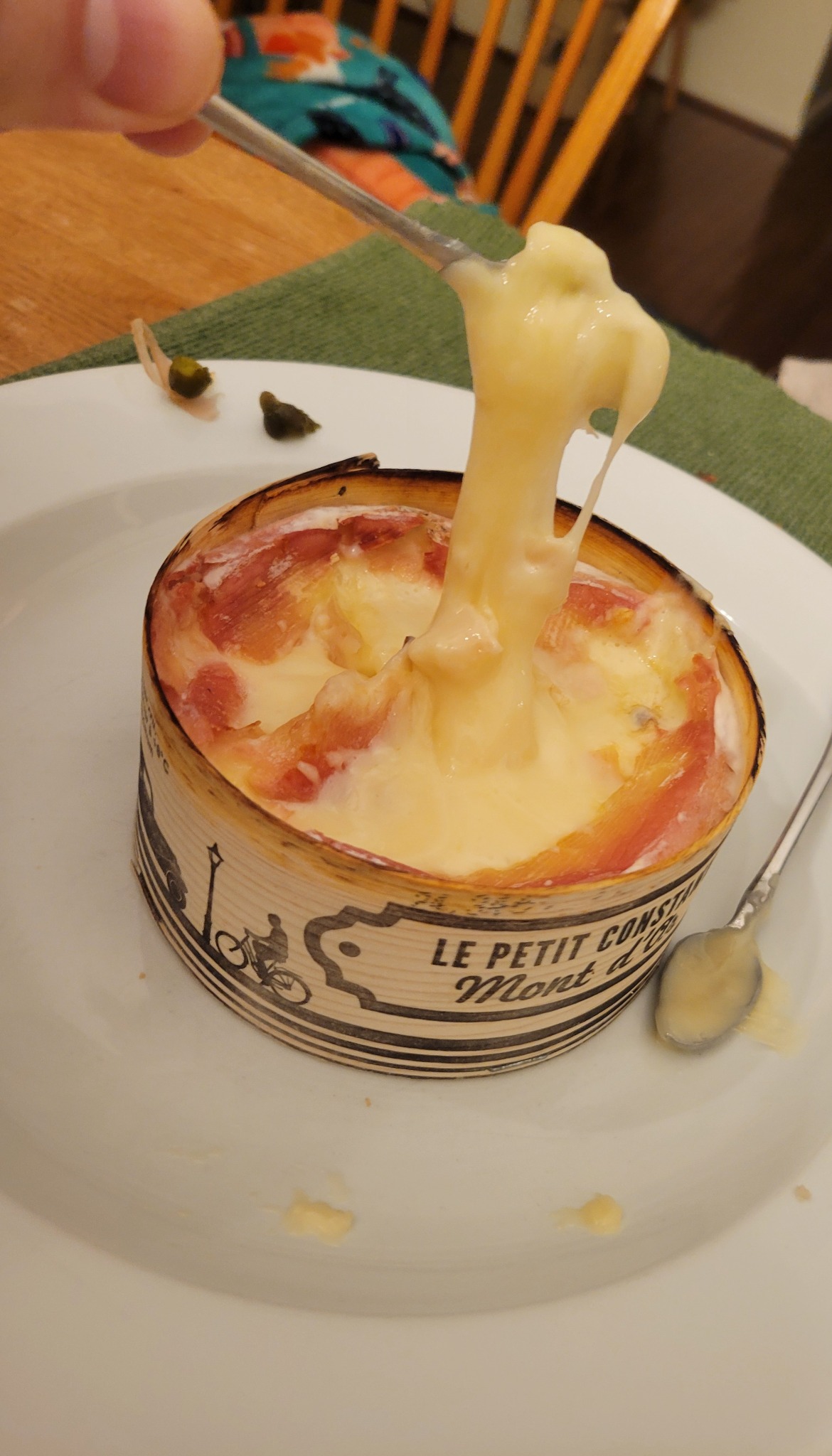 Spoon lifting molten Mont d’Or while the original printing remains visible on the wooden package